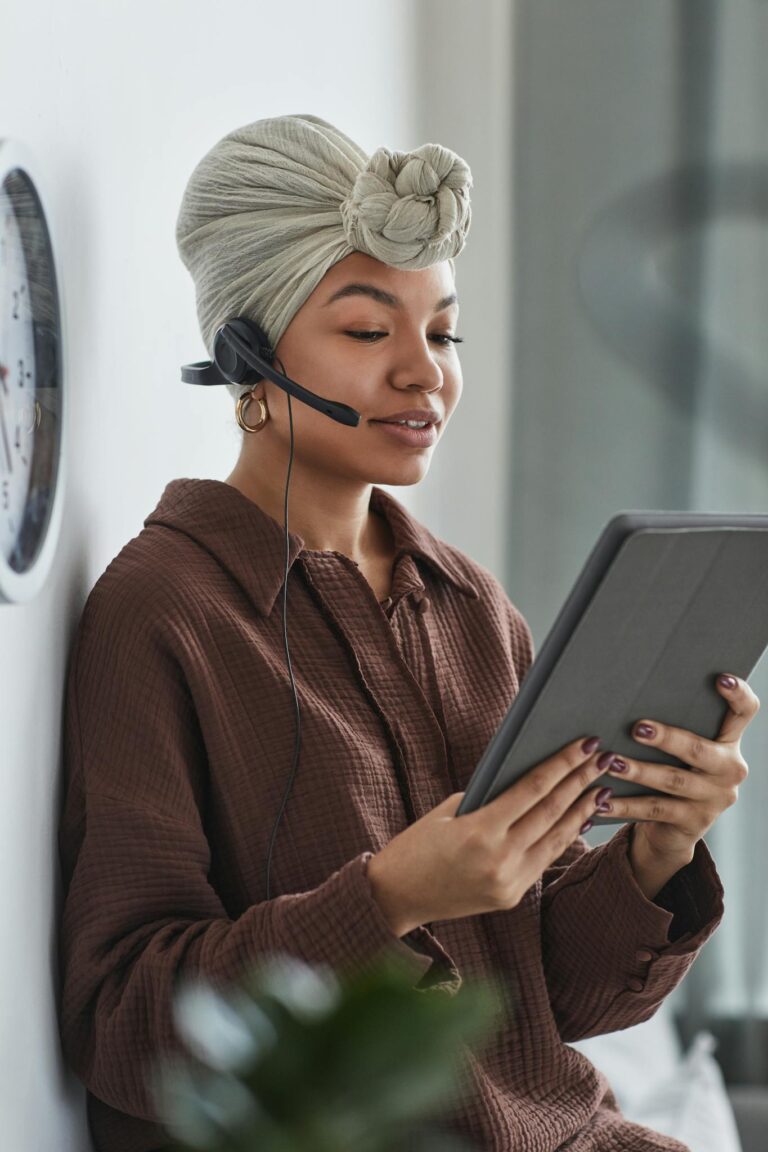 Content African American female call agent with microphone reading information on tablet while working in room with blurred green plants